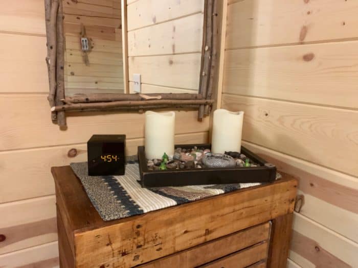 Wooden drawers with a rock and 2 candle display along with a black cube digital clock and mirror hanging on wall