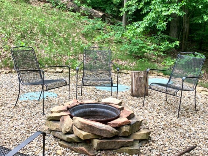 Black steel chairs around a fire pit made from piled rocks in front of green trees