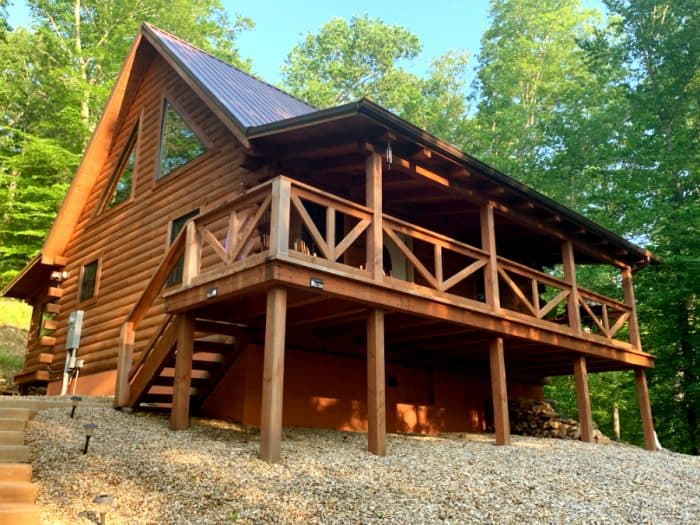 Log cabin with front deck and gravel at the bottom sitting among tall green trees with a blue sky