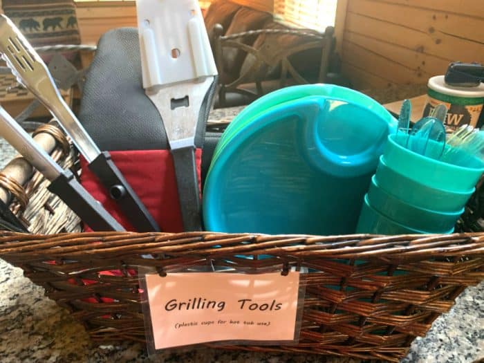 Brown woven oblong basket with a sign saying 'grilling tools' holding grilling tongs, hamburger flipper and blue plastic plates, cups and cutlery.