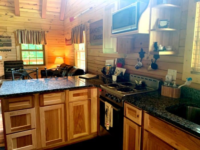 Kitchen area of a log cabin with wooden cabinets and granite countertops, a stove and coffee cups hanging on the wall