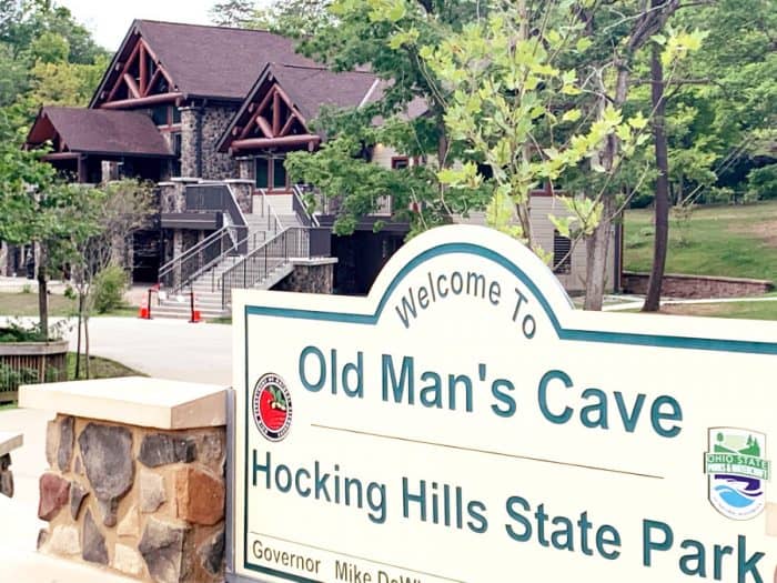 Stone and wooden 2 story building in background with a sign in front reading Welcome to Old Man's Cave Hocking Hills State Park