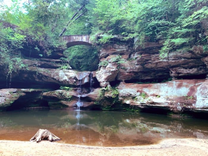 Rock structure supporting a small waterfall into a pond below it with green trees all around.