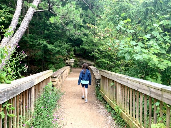 Young girl walking across a wooden and stone bridge on a dirt trail in a forest